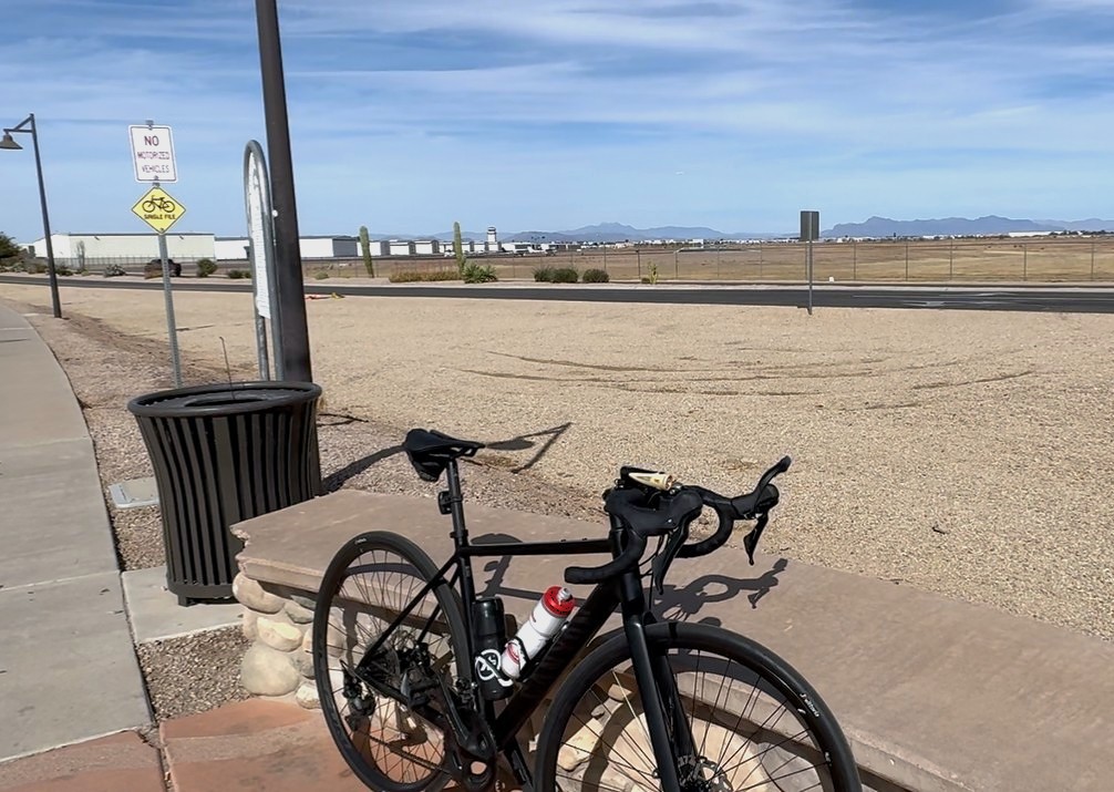 A bicycle leans against a bench in front of a fenced airport runway.
