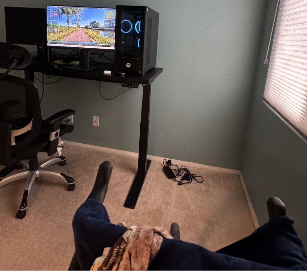 A bicycle sits on an indoor trainer facing a computer on a desk. 