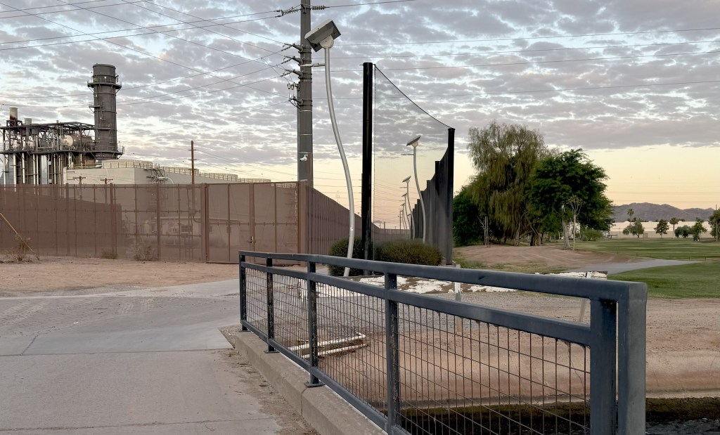 A pathway over a canal between a golf course and a power plant. 