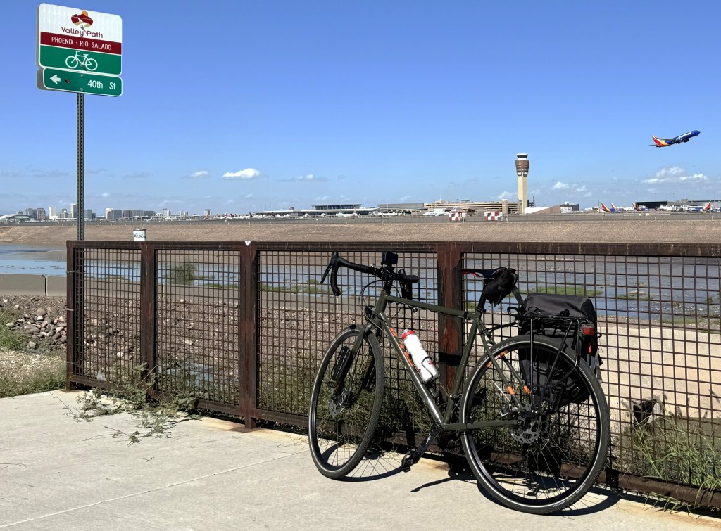 A bicycle leans against a fence with an airplane taking off in the background. 