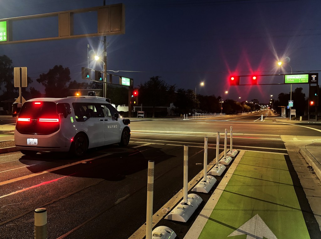 A Waymo self-driving car waits at a stop light next to a protected bicycle lane.